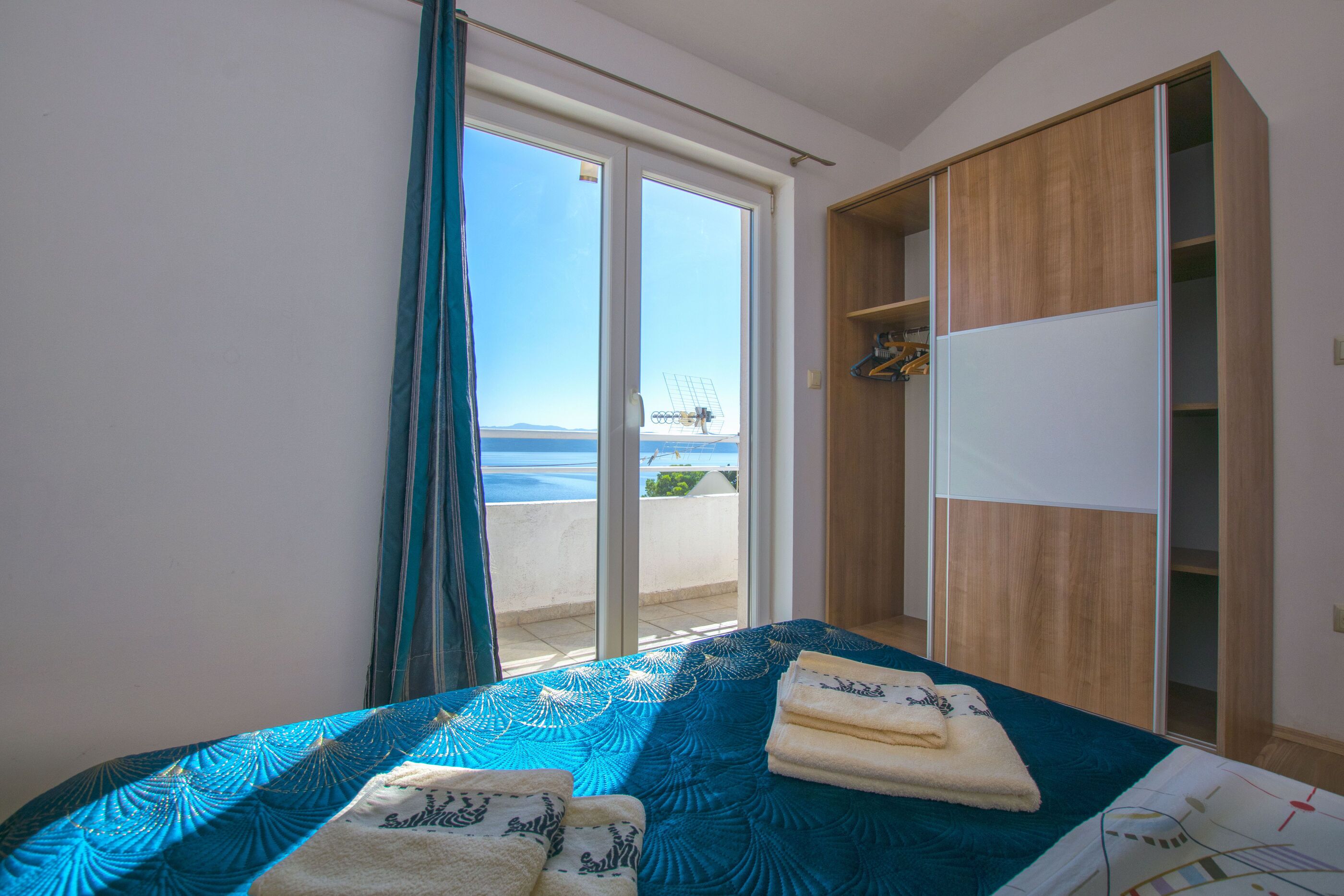 Une chambre à coucher comprenant un lit, une armoire en bois et une porte-fenêtre coulissante donnant sur un balcon avec vue sur la mer.
