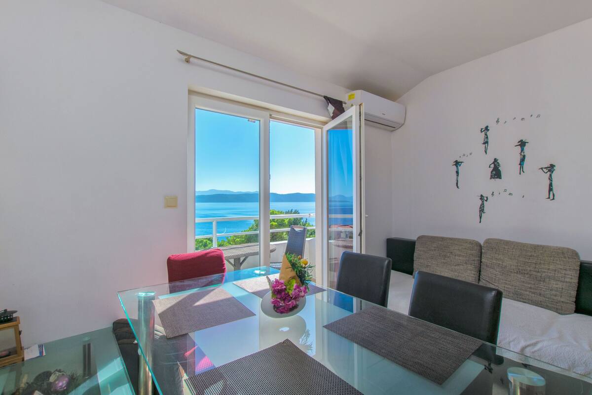 Uma sala de jantar moderna com mesa de vidro, cadeiras e vista para o mar através de grandes janelas.