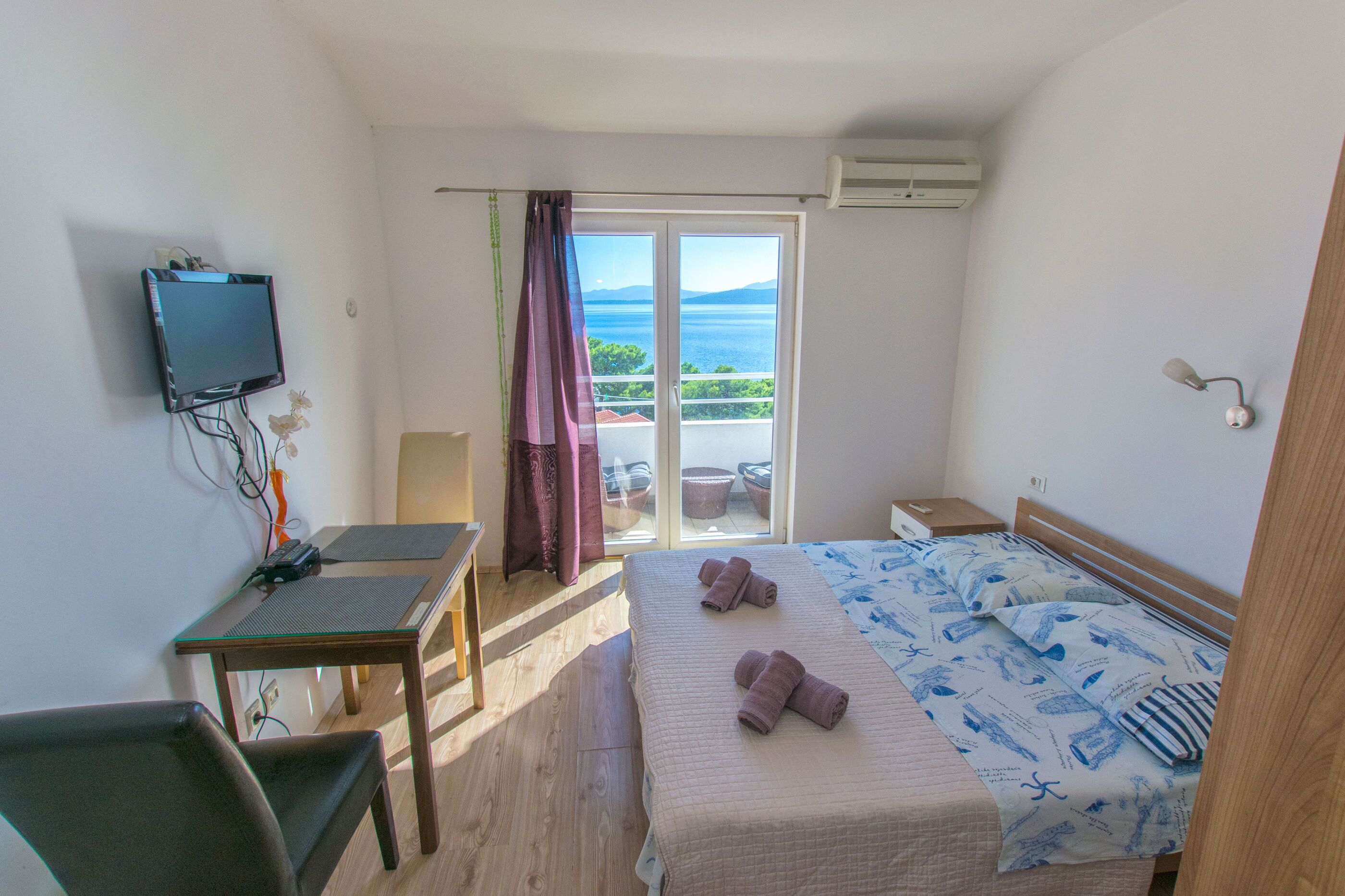 Een slaapkamer met een bed, een bureau, een stoel, een televisie en een balkon met uitzicht op zee.