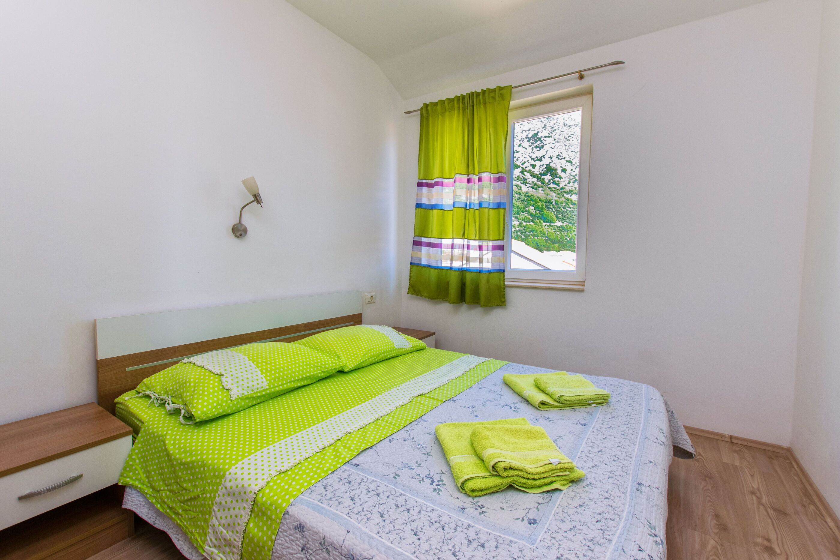 A bedroom with a bed, a nightstand, a window with curtains, and a wall-mounted lamp.