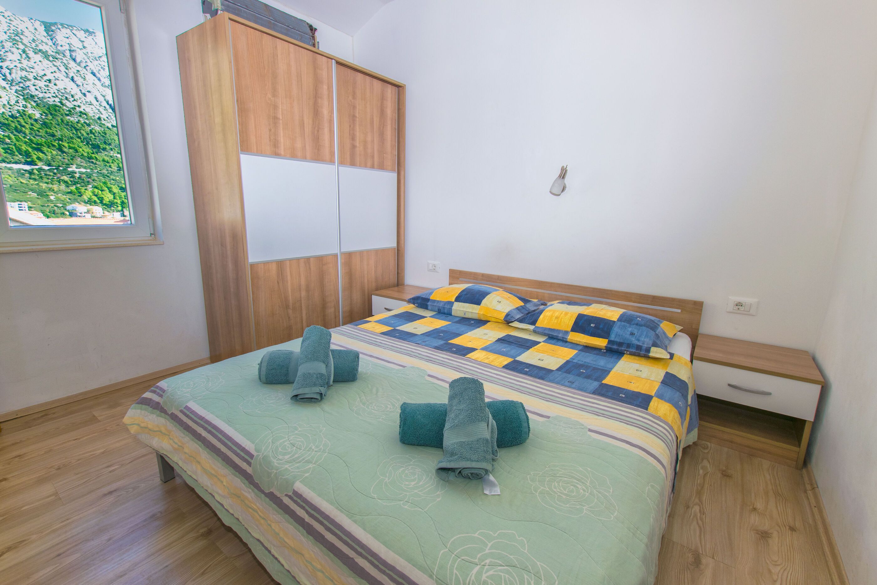 Ein Schlafzimmer mit Bett, Kleiderschrank, Nachttisch und einem Fenster mit Blick ins Grüne.