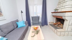 A living room with a grey sofa, a blue armchair, a wooden coffee table with fruits and a branch, and a stone fireplace with logs. - House Rosemary (Slivno)