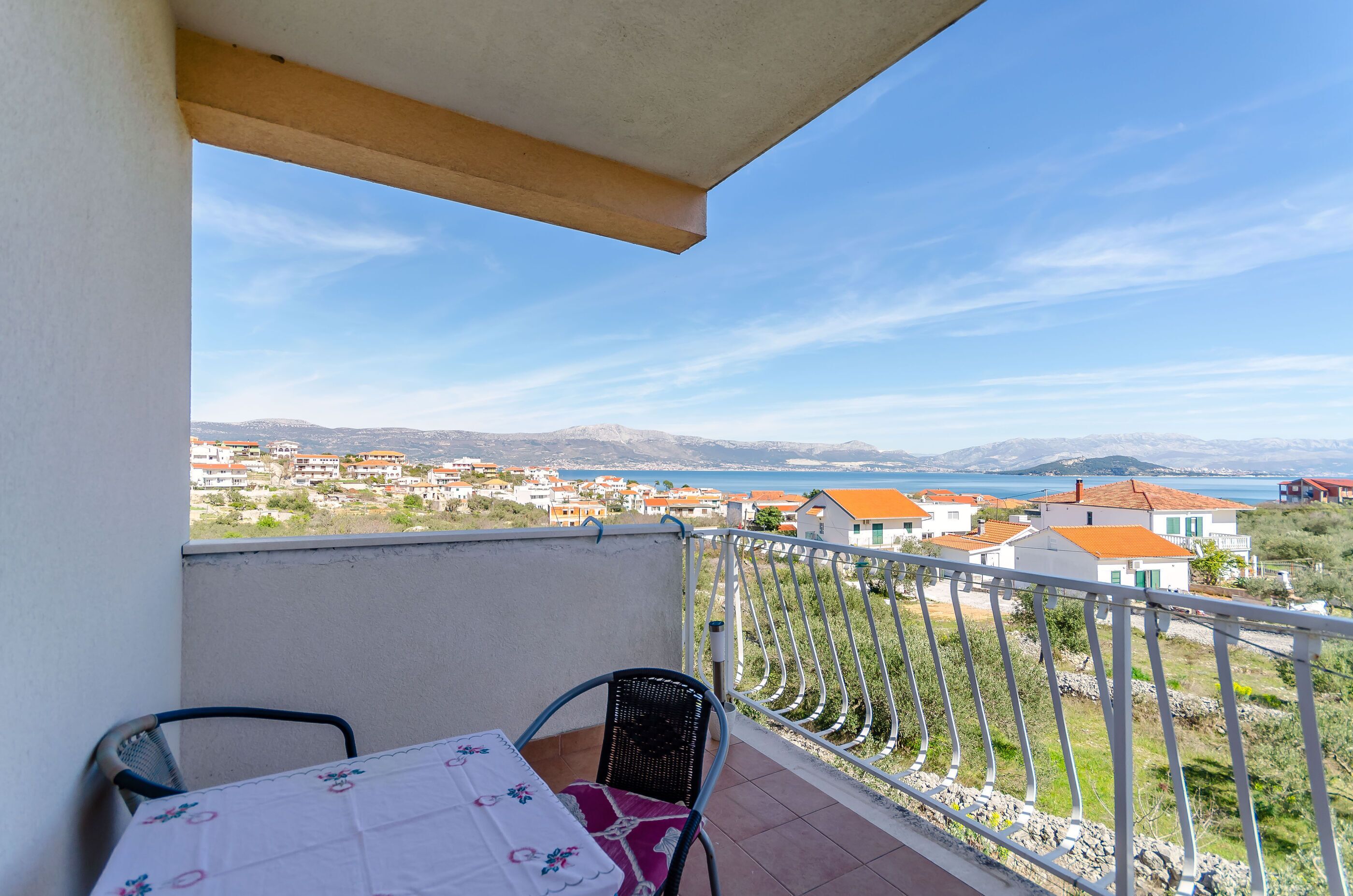 A balcony with a table and chairs, overlooking a coastal town with houses and a sea view.