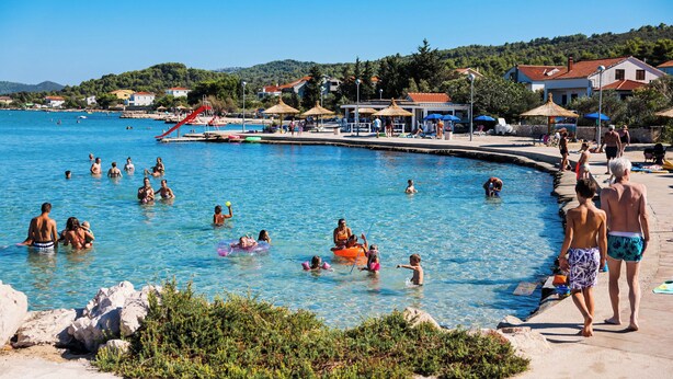 A swimming pool area with people swimming and sunbathing, surrounded by houses and greenery. - House Sea Stone (Pasman)