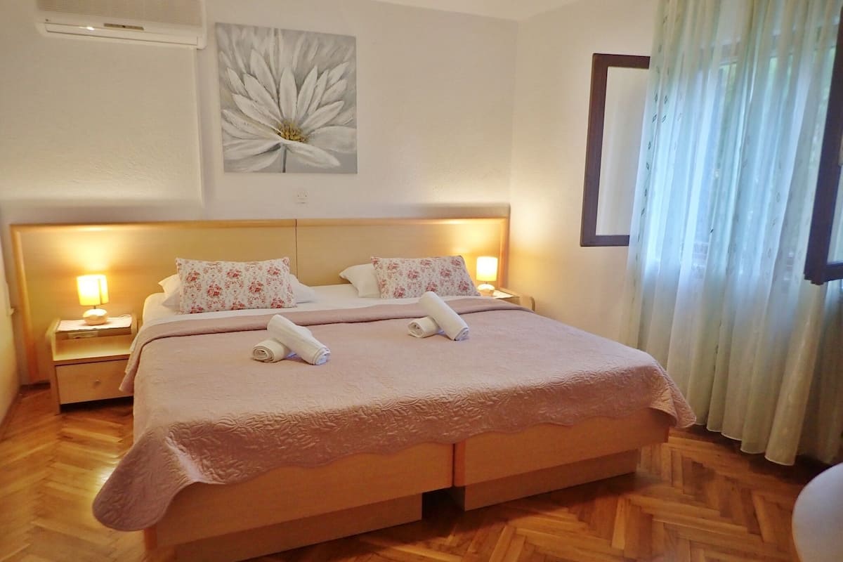 A hotel room with a large bed, two bedside tables with lamps, a floral painting on the wall, and a window with curtains.