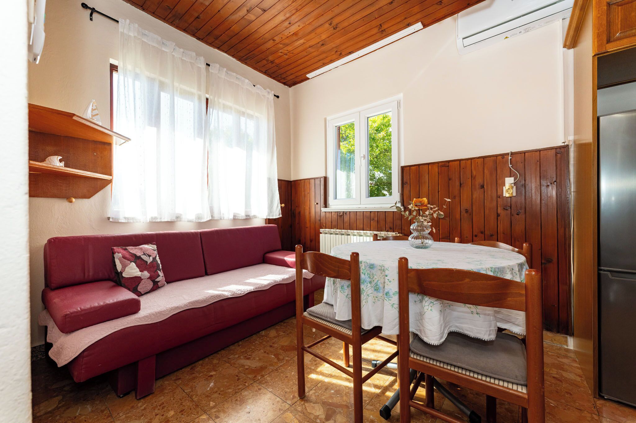 A cozy room with a wooden ceiling, a red sofa, a dining table with chairs, a window with a view of greenery, and a built-in refrigerator.