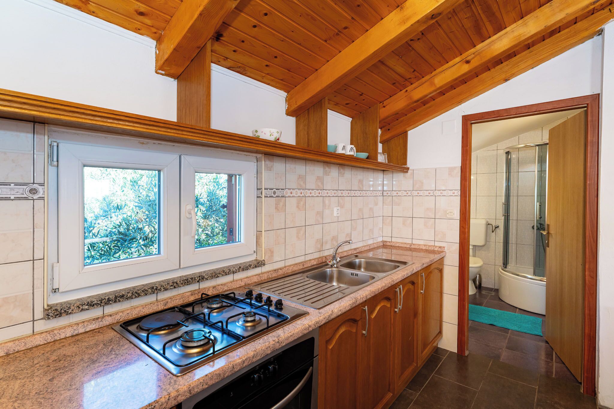 A kitchen with a gas stove, double sink, wooden cabinets, and a window with a view of greenery.