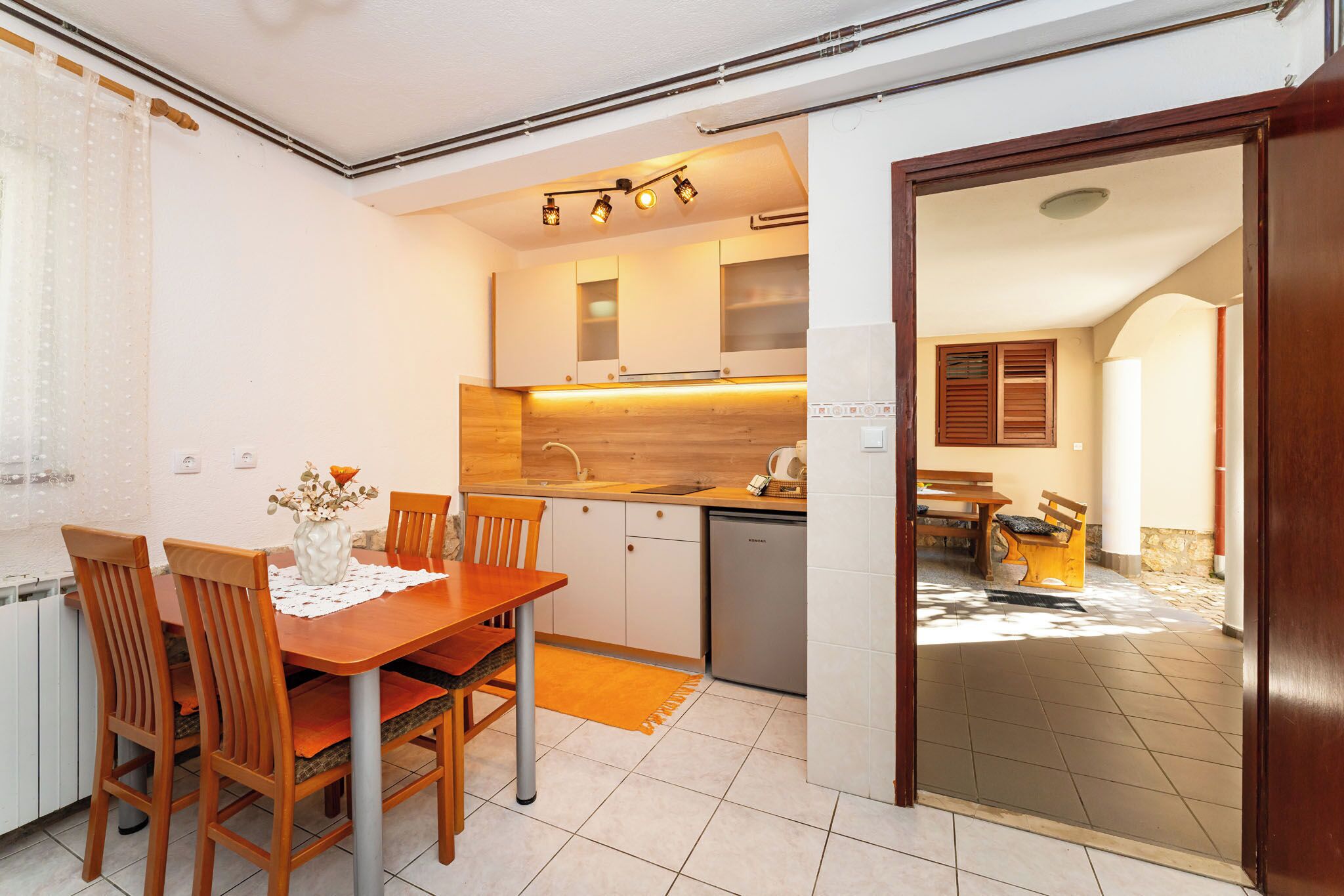 Una cocina compacta con armarios de madera, horno empotrado y lavavajillas. Cuenta con un comedor con mesa y sillas de madera, y un pequeño espacio exterior con banco y mesa.