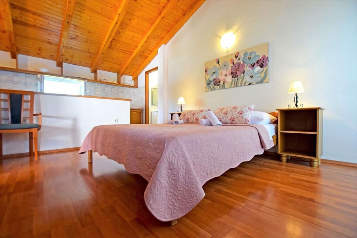 A bedroom with a wooden ceiling, a bed with a pink quilt, a painting on the wall, and a nightstand with a lamp.