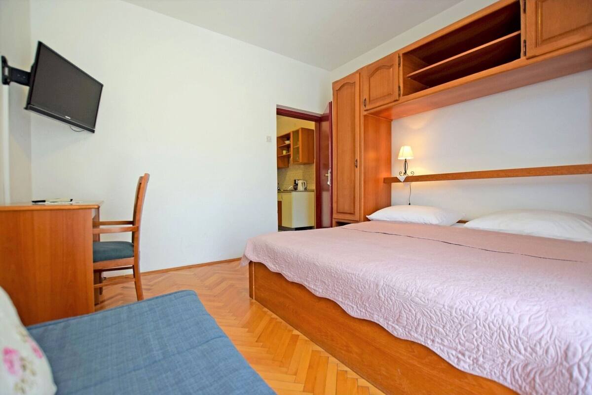 A bedroom with a bed, a desk, a chair, a television, and a kitchen area visible through an open door.