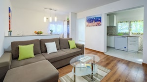 Ein modernes Wohnzimmer mit einem braunen Ecksofa, einem Glastisch und einer Küche im Hintergrund.