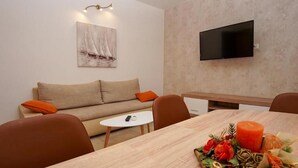 A modern living room with a sofa, a dining table with a centerpiece, a small white table, and a mounted TV. - Apartment Branka (Vodice)