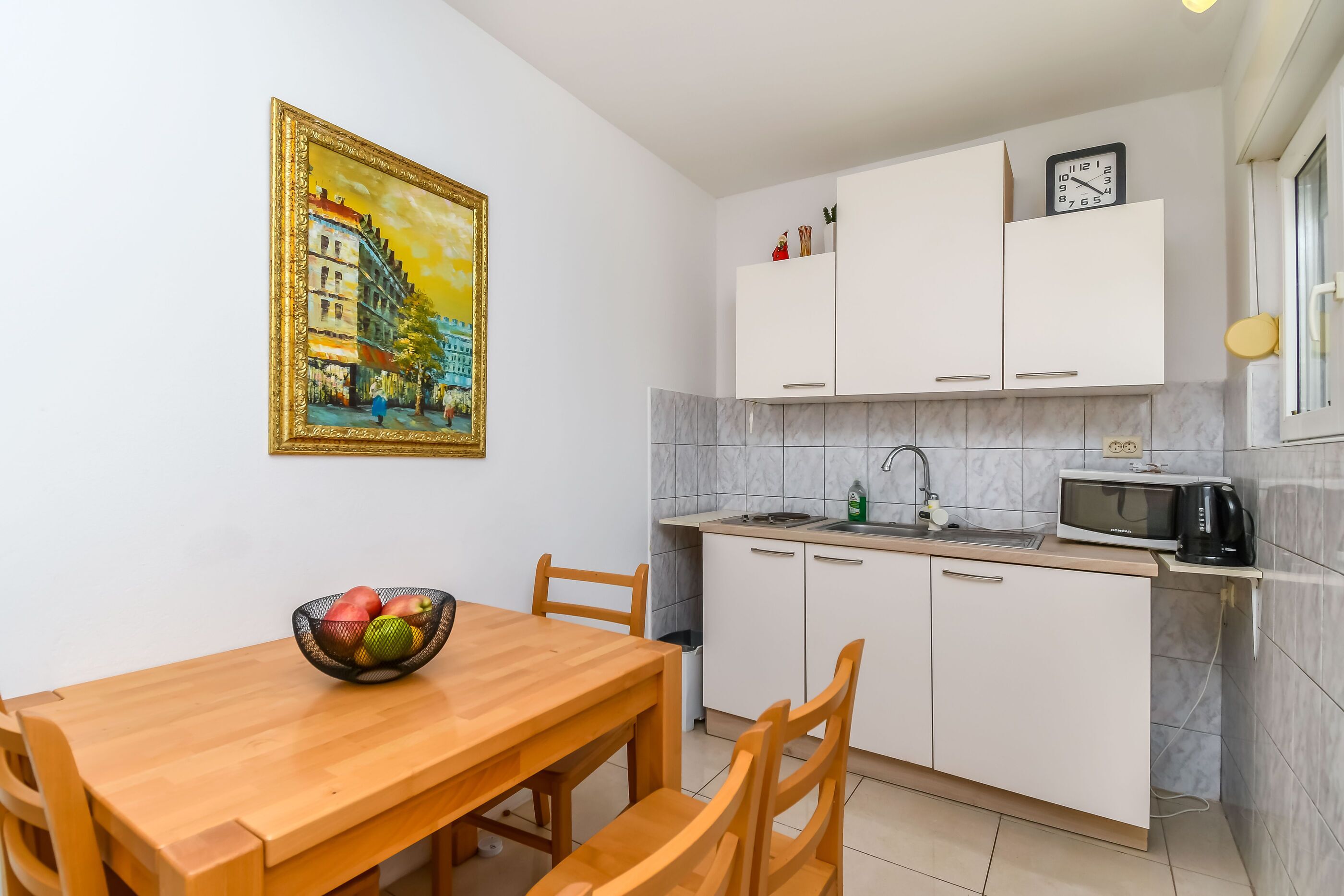 A kitchen with white cabinets, a microwave, and a sink. There is a painting on the wall and a bowl of fruit on the table.