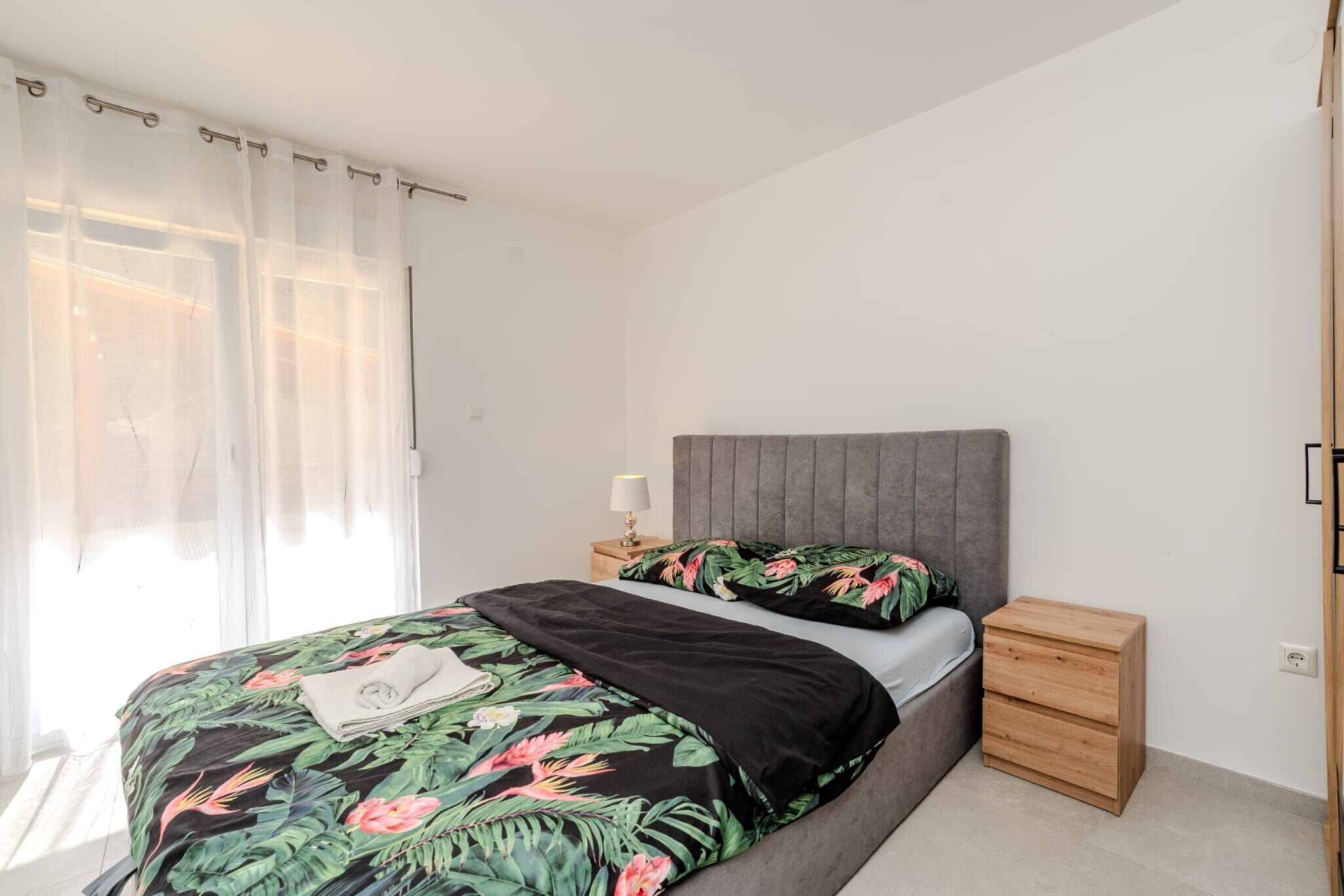 Une chambre à coucher comprenant un lit, une table de chevet, une commode en bois et des rideaux légers.