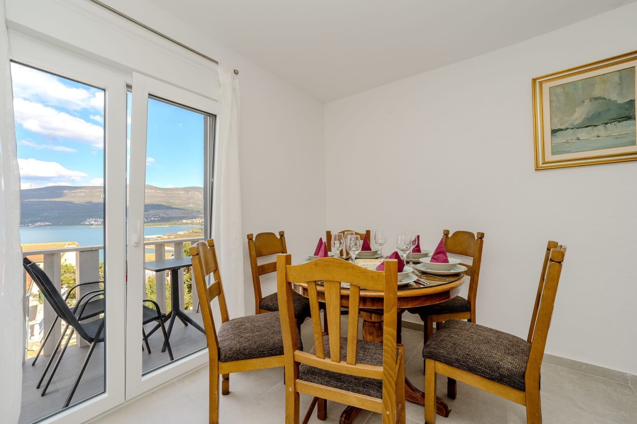 A dining area with a table set for four, wooden chairs, and a view of a body of water through large windows.
