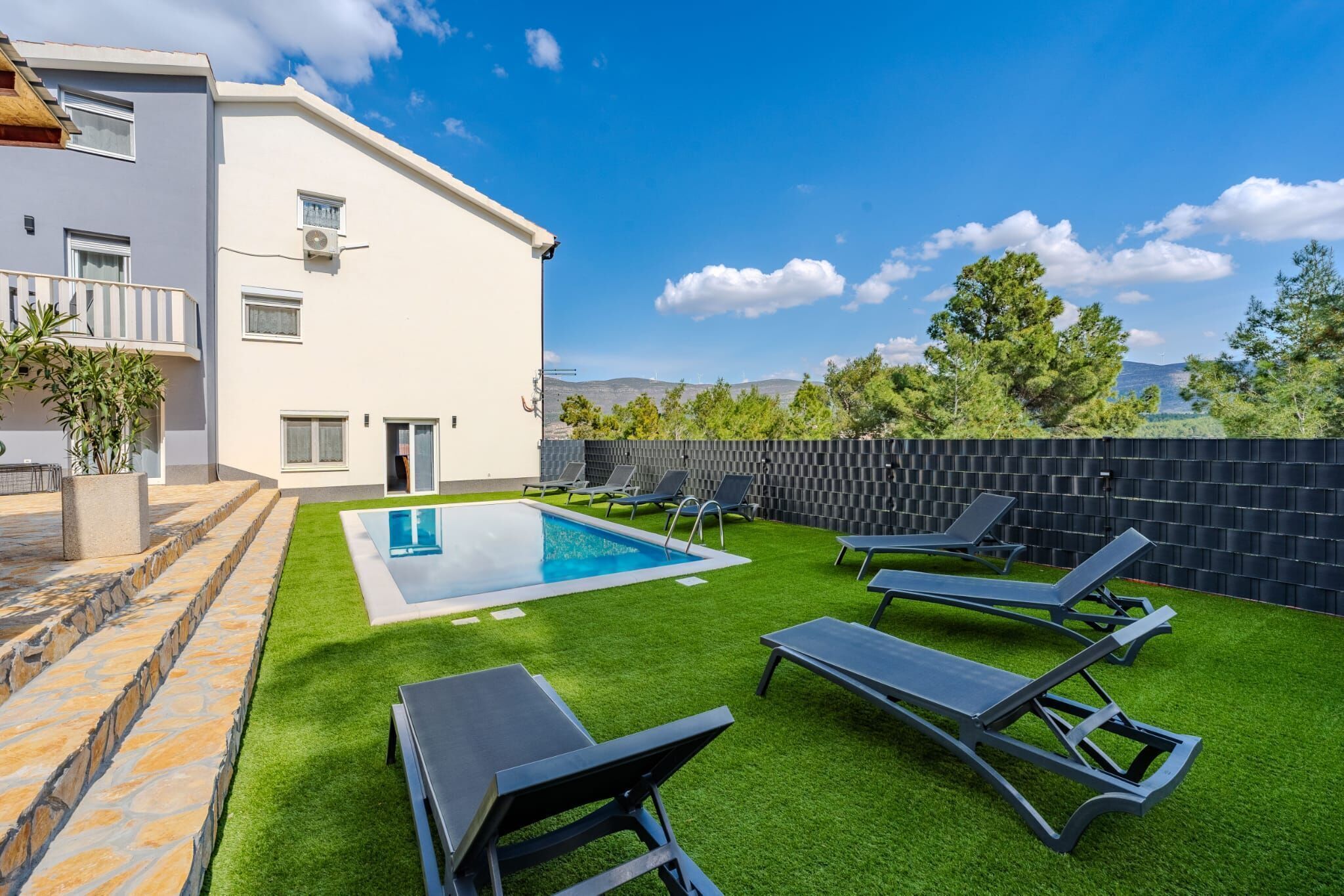 A modern outdoor pool area with artificial grass, lounge chairs, and a stone pathway leading to a building with a white facade and a door.