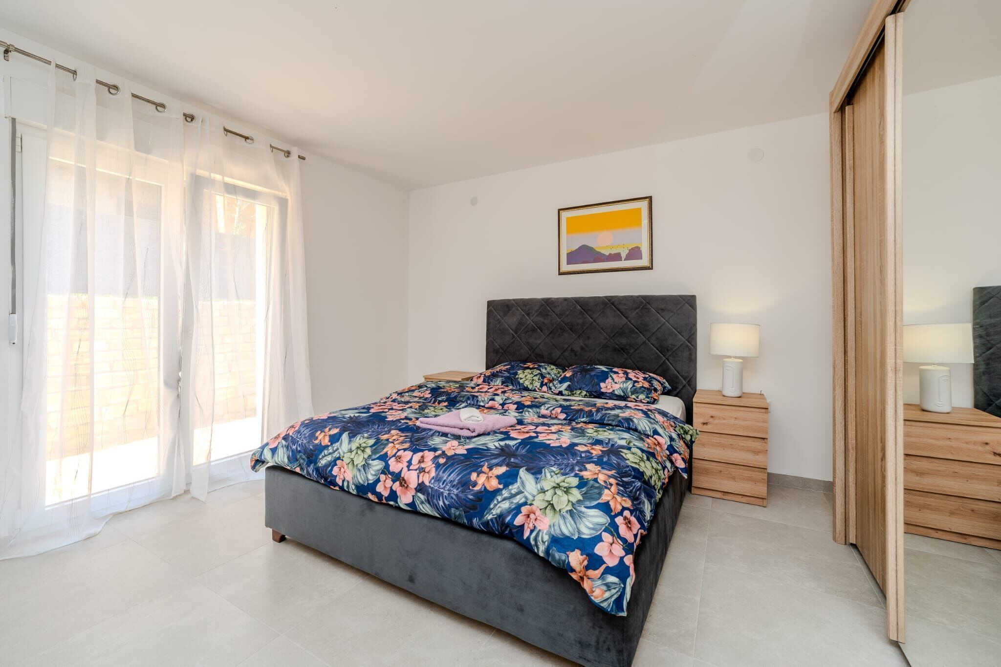A bedroom with a bed, two bedside tables, a nightstand, a wardrobe, and a window with sheer curtains.