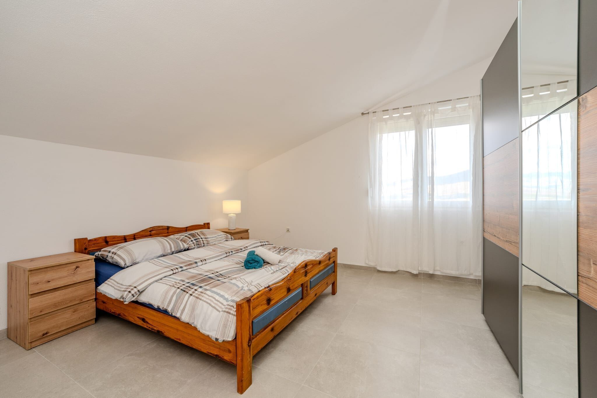 A bedroom with a wooden bed, a nightstand, a drawer unit, and a large mirror.
