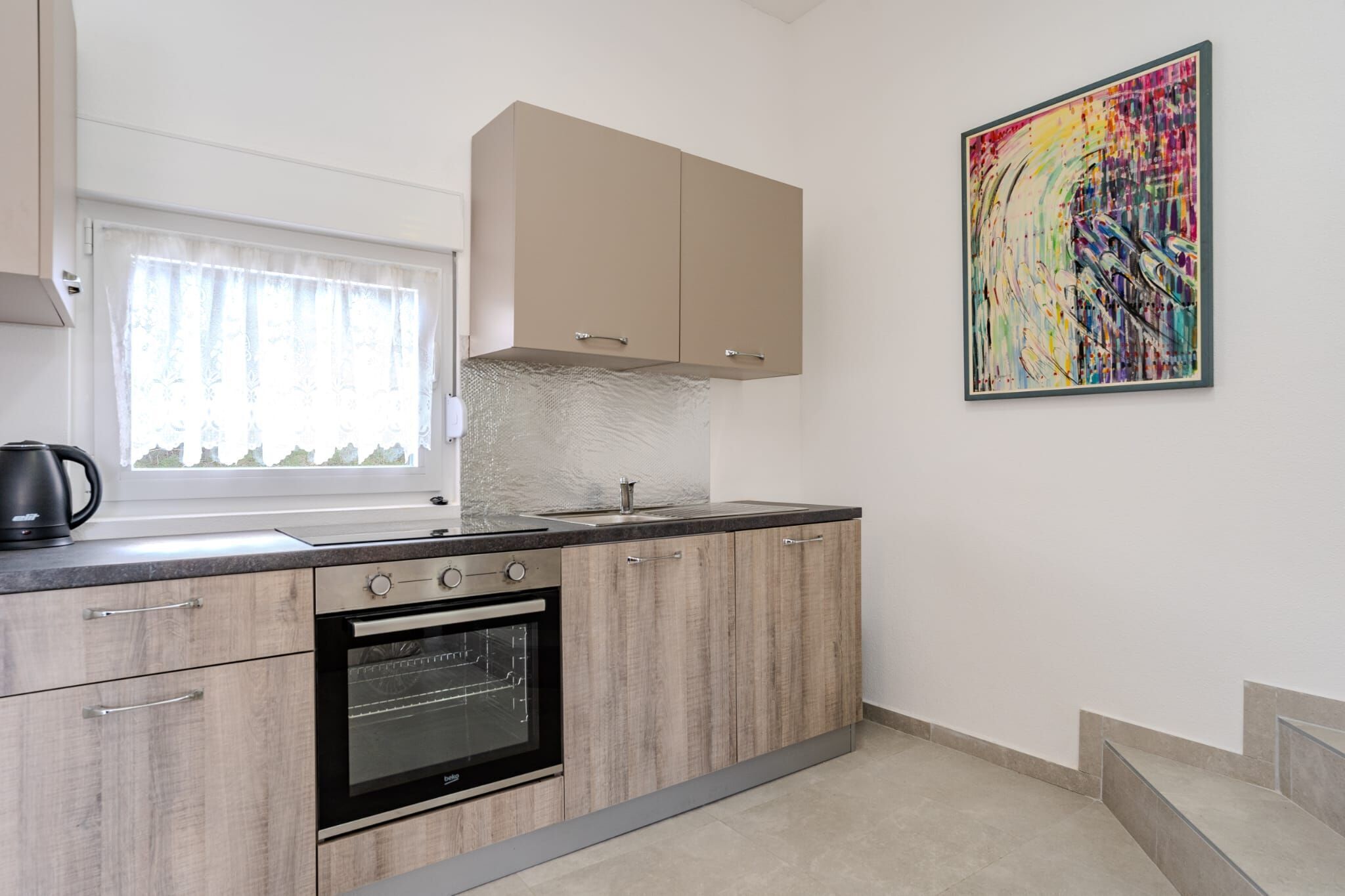 A modern kitchen with light wood cabinets, a stainless steel oven, and a large window with sheer curtains.