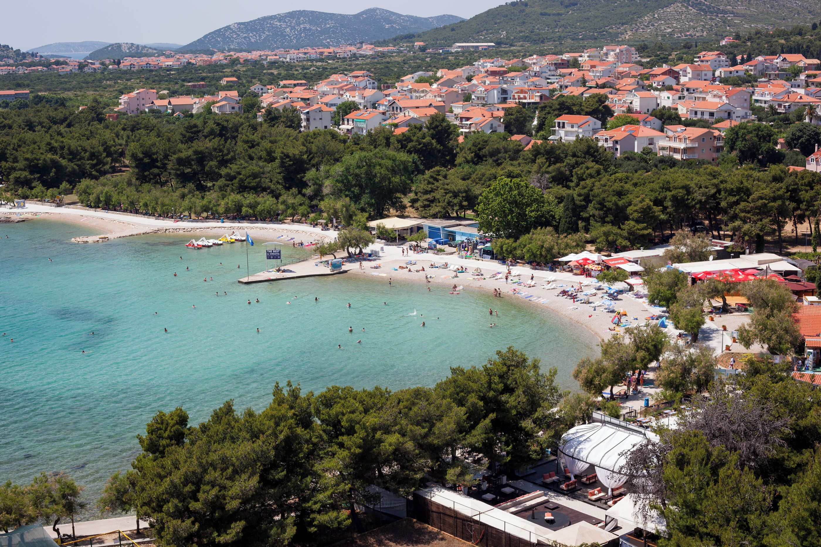 Eine Küstenstadt mit Strand, klarem türkisblauem Wasser und roten Dächern.