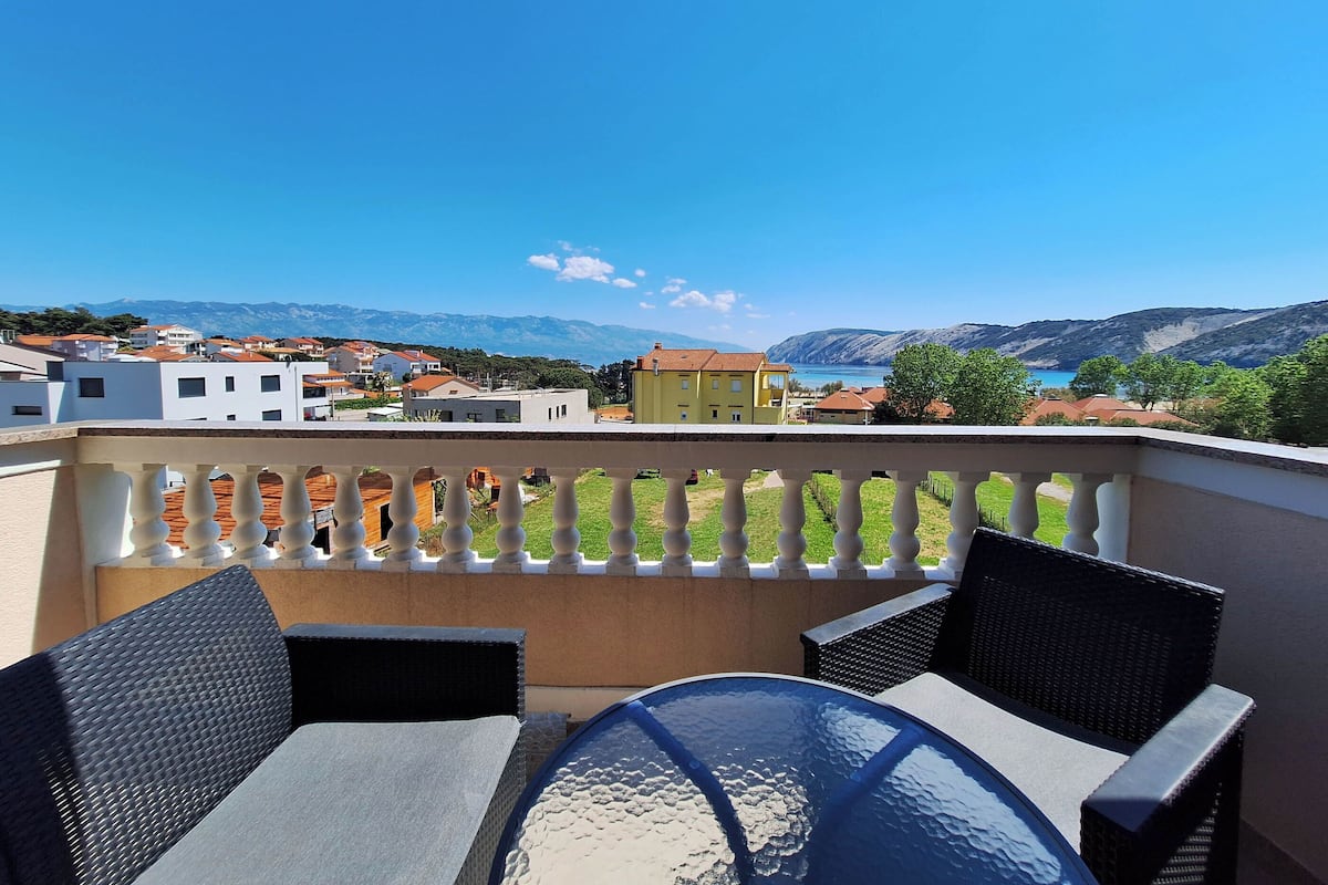 A balcony with wicker furniture and a glass table, overlooking a town with buildings and a lake.