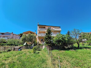 Monikerroksinen asuintalo, jossa on parvekkeet, ja jonka ympärillä on vehreää kasvillisuutta ja kirkas, sininen taivas.