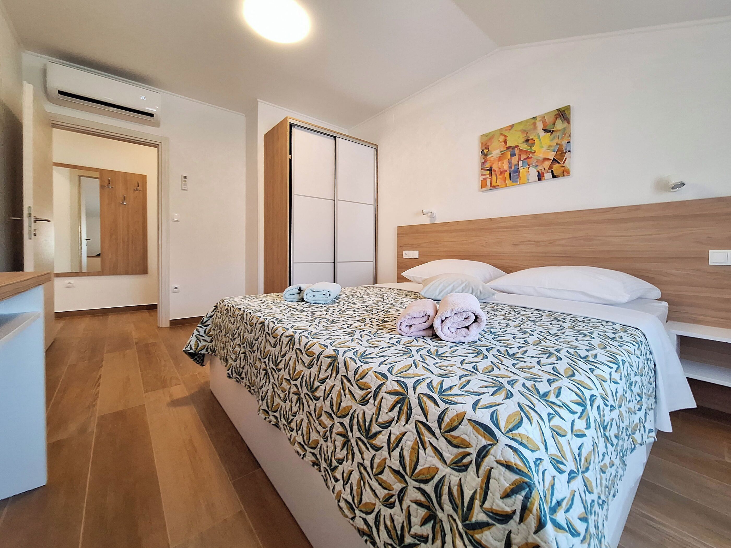 Habitación de hotel con cama, cabecera de madera, un cuadro en la pared y baño con espejo y puerta.