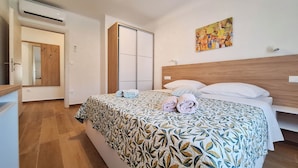 A hotel room with a bed, a wooden headboard, a painting on the wall, and a bathroom with a mirror and door. - Apartments Dijana (Lopar)