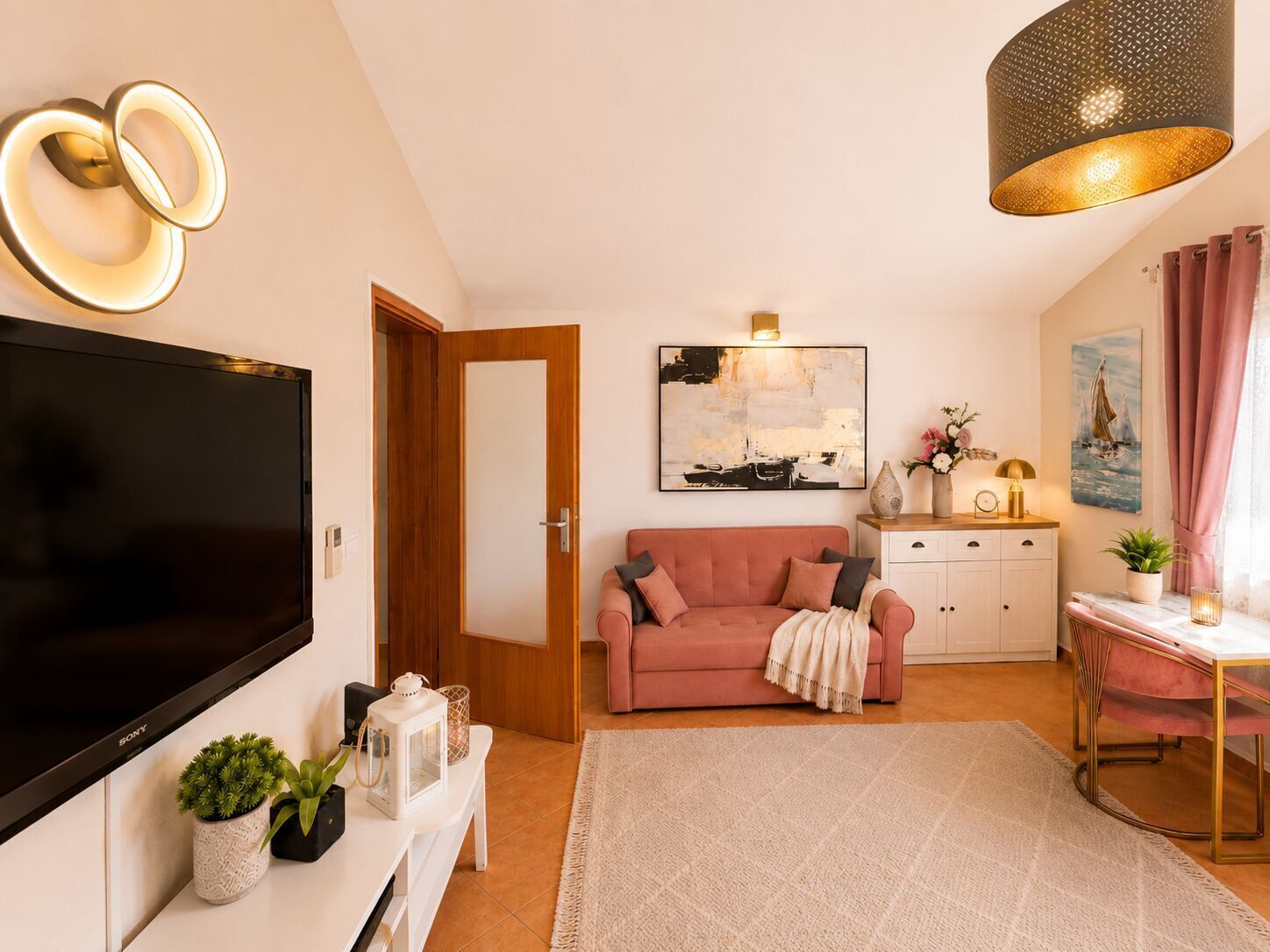 A modern living room with a flat-screen TV, a pink sofa, a white cabinet, a dining table with a gold frame, and a large abstract painting on the wall.