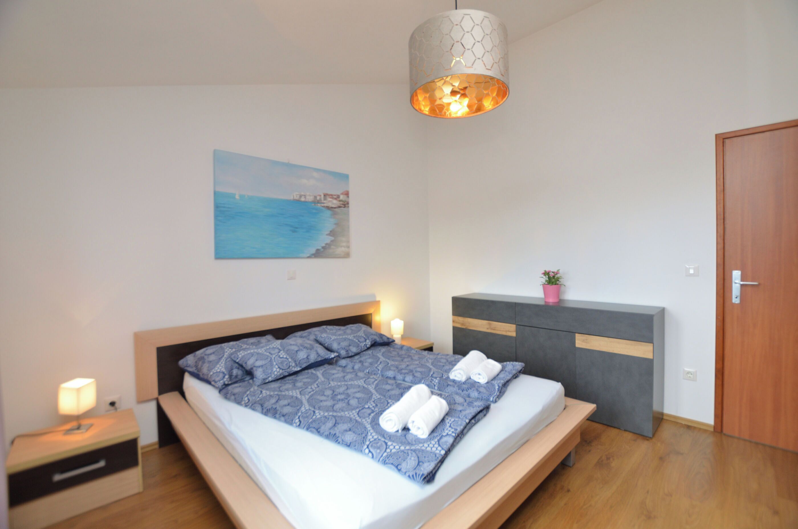 A modern bedroom with a bed, bedside tables, a nightstand, a dresser, a painting, and a hanging light fixture.