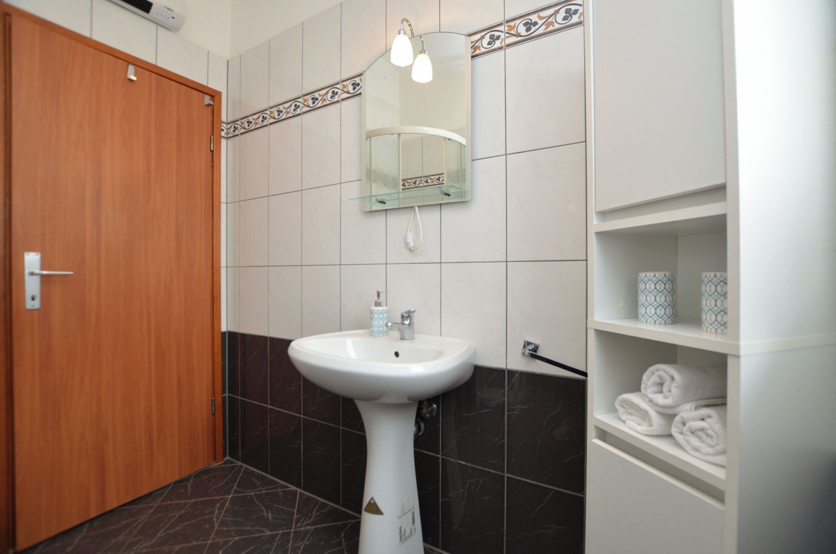 Un baño con puerta de madera, un lavamanos pedestal, un espejo con iluminación y un estante con toallas.