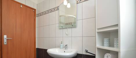 Un baño con puerta de madera, un lavamanos pedestal, un espejo con iluminación y un estante con toallas.