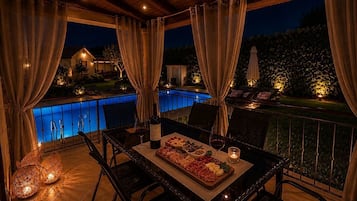 Área de refeições ao ar livre com vista para a piscina, teto de madeira e mesa posta com comida e bebidas.