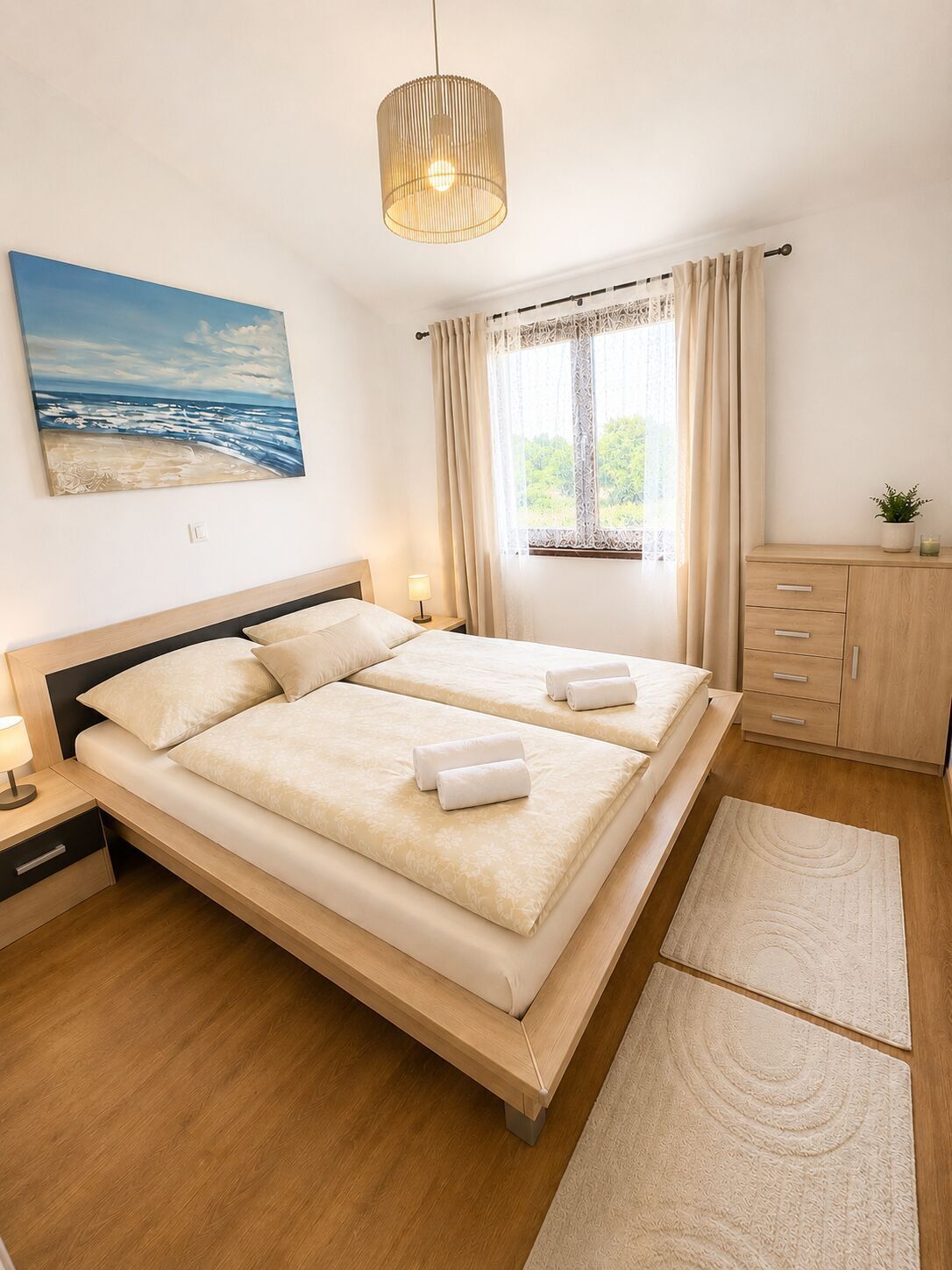 Uma cama de casal com duas almofadas, uma mesa de cabeceira com abajur, uma janela com cortinas, um quadro de uma praia e um armário de madeira.