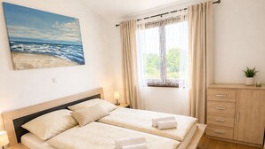 Een tweepersoonsbed met twee kussens, een nachtkastje met een lamp, een raam met gordijnen, een schilderij van een strand en een houten kast.