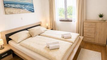 Uma cama de casal com duas almofadas, uma mesa de cabeceira com abajur, uma janela com cortinas, um quadro de uma praia e um armário de madeira.