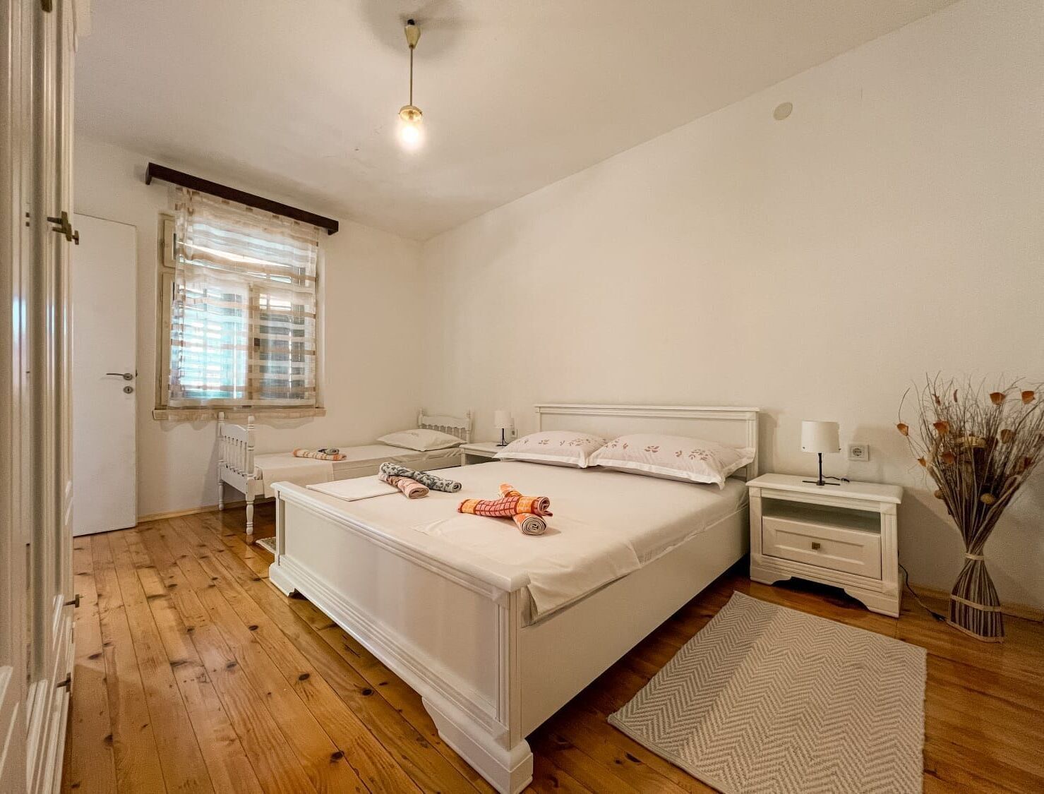 Ein Schlafzimmer mit einem großen Bett, zwei Nachttischen, einem Schreibtisch und einem Fenster mit Jalousien. Der Boden ist aus Holz.