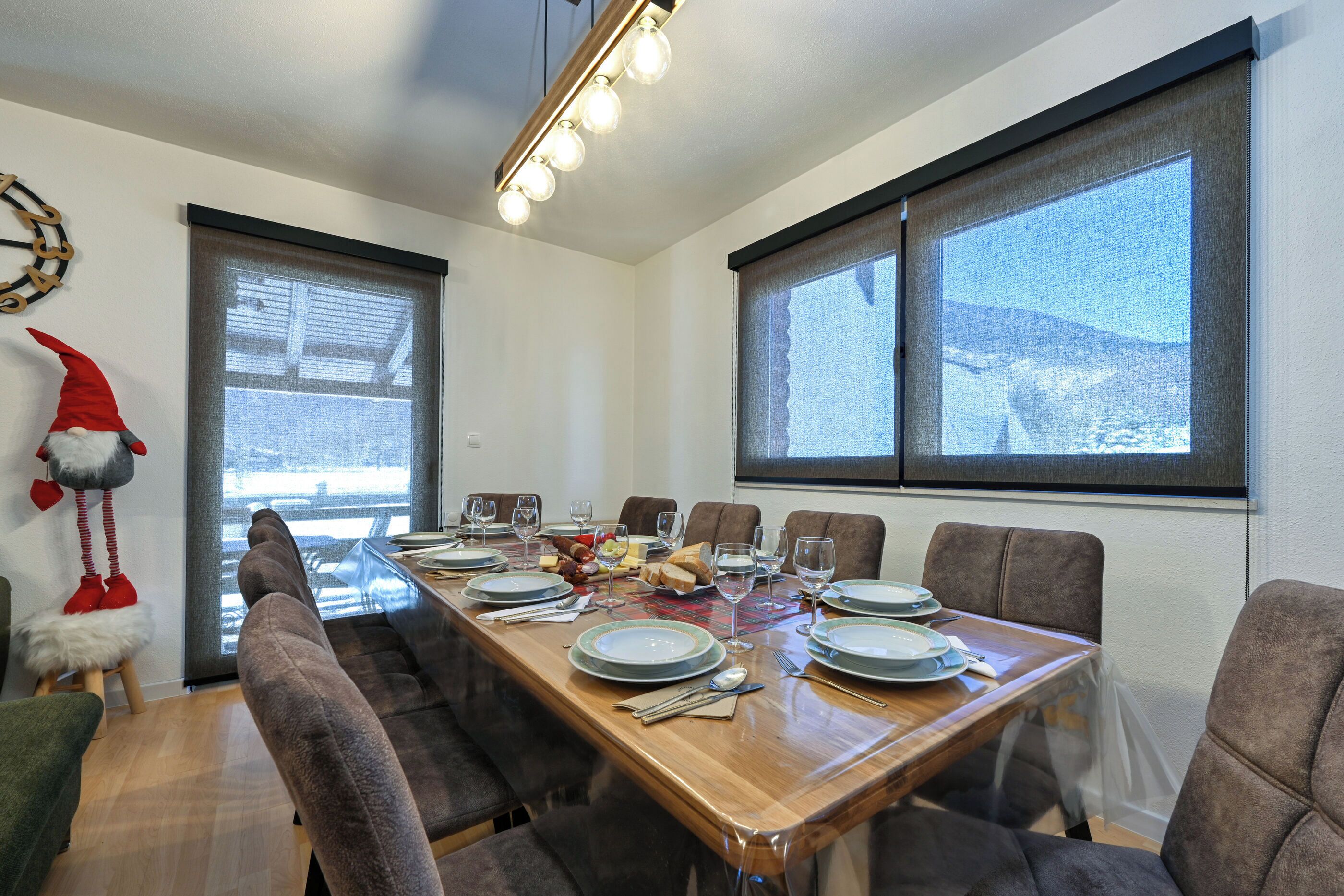 A dining room with a wooden table set for six, surrounded by chairs, with a large window and a decorative wall piece.