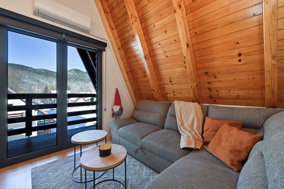 Ein gemütliches Zimmer mit einem grauen Sofa, einem kleinen runden Tisch und einem großen Fenster mit Blick auf eine verschneite Landschaft.