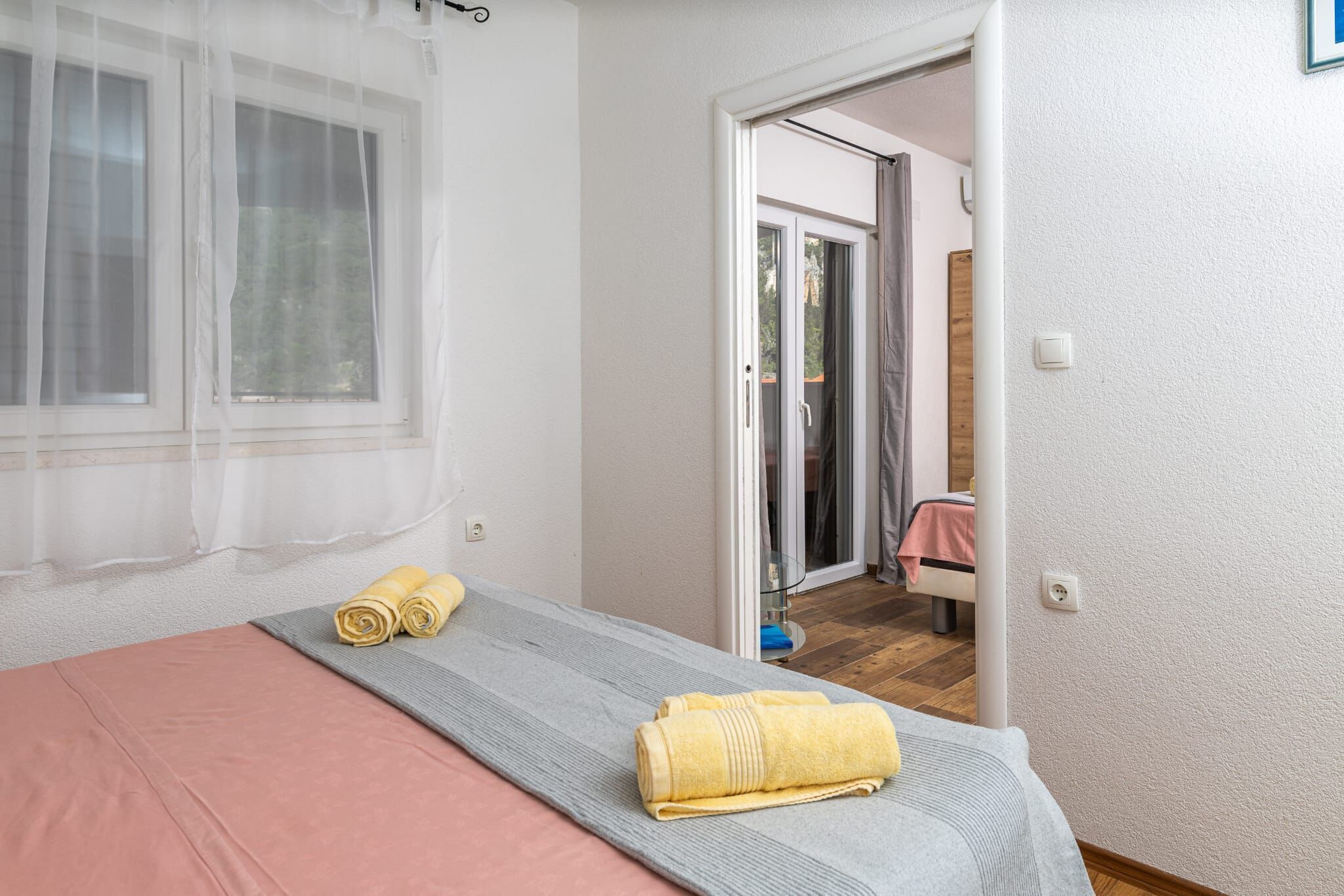 Ein Schlafzimmer mit einem Bett, einem Fenster mit durchsichtigen Vorhängen, einer Tür, die auf einen Balkon führt, und einem kleinen Tisch mit Handtüchern.