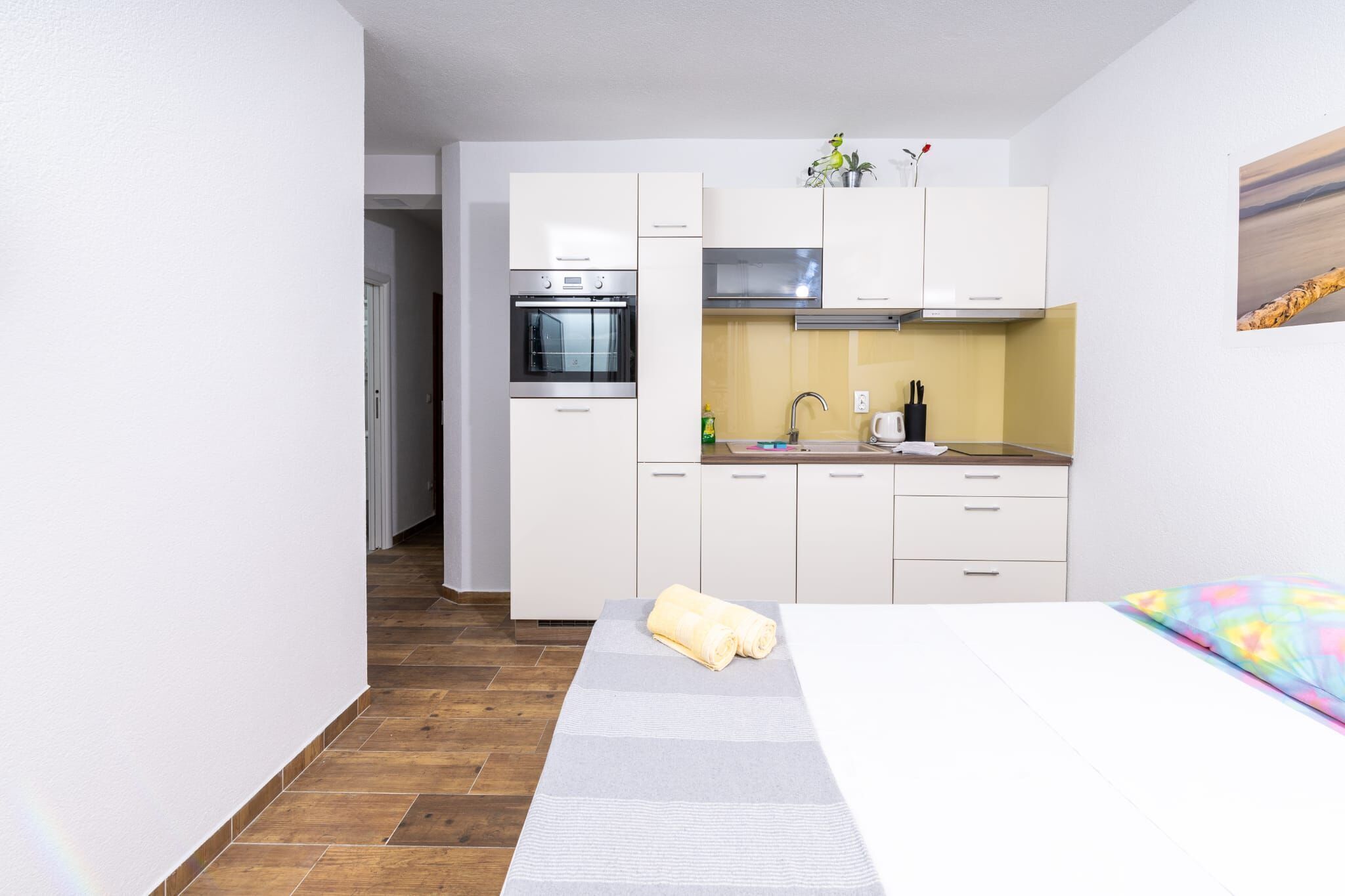 A compact hotel room with a kitchenette, a bed with a colorful pillow, and a wooden floor.