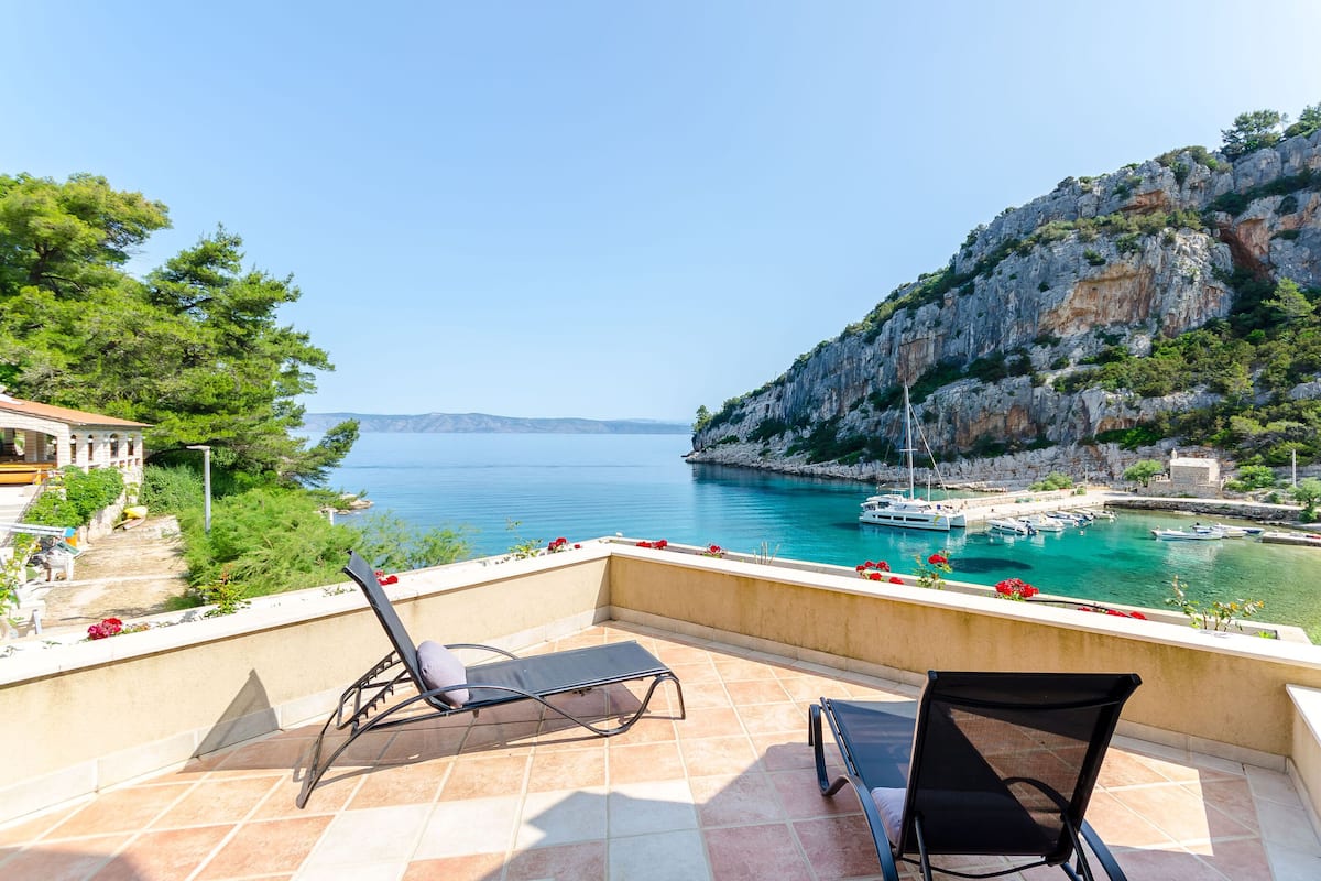 A balcony with two lounge chairs overlooking a coastal marina with boats and a rocky cliff.