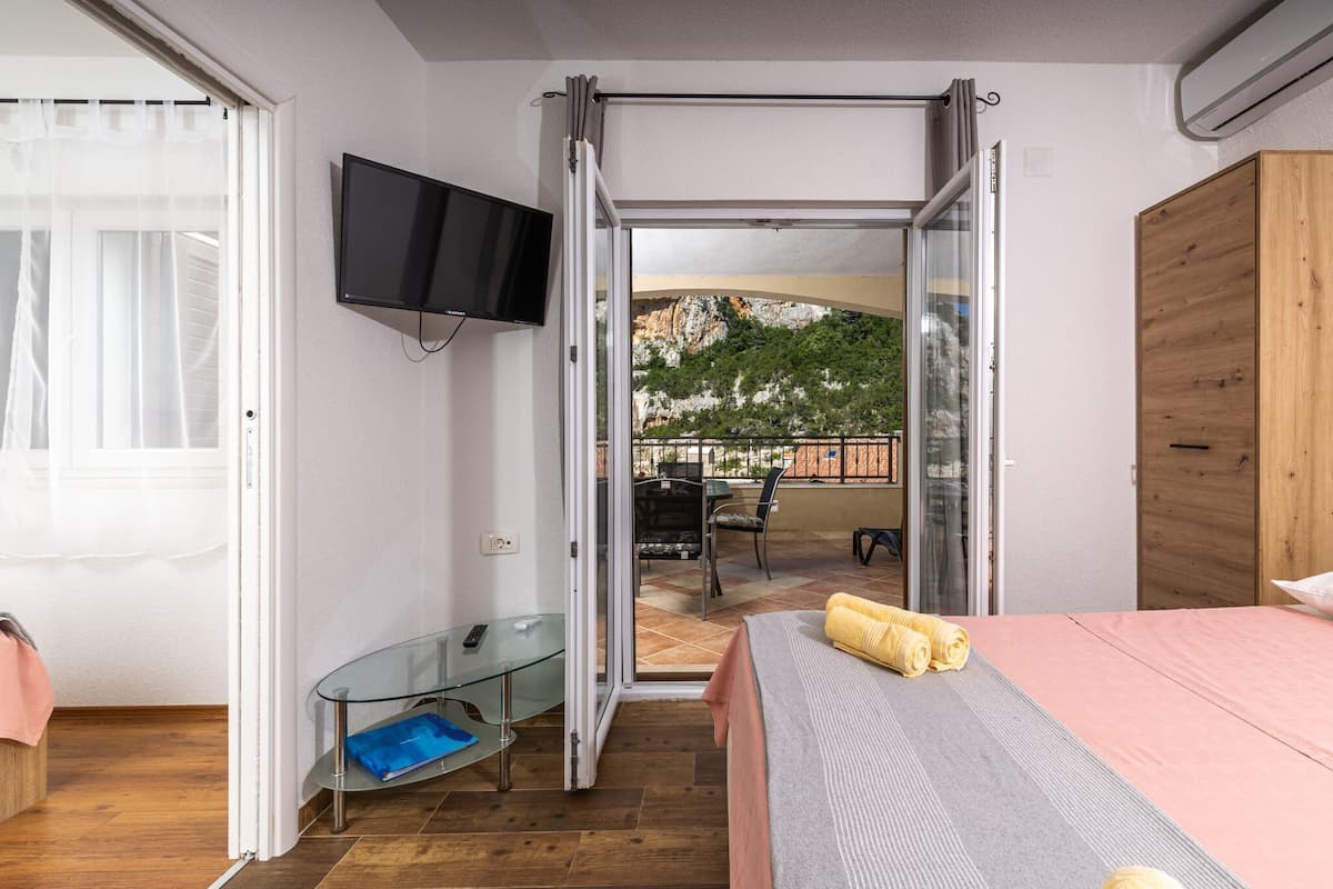 A bedroom with a bed, a TV, a glass table, and a sliding door leading to an outdoor patio with a table and chairs.