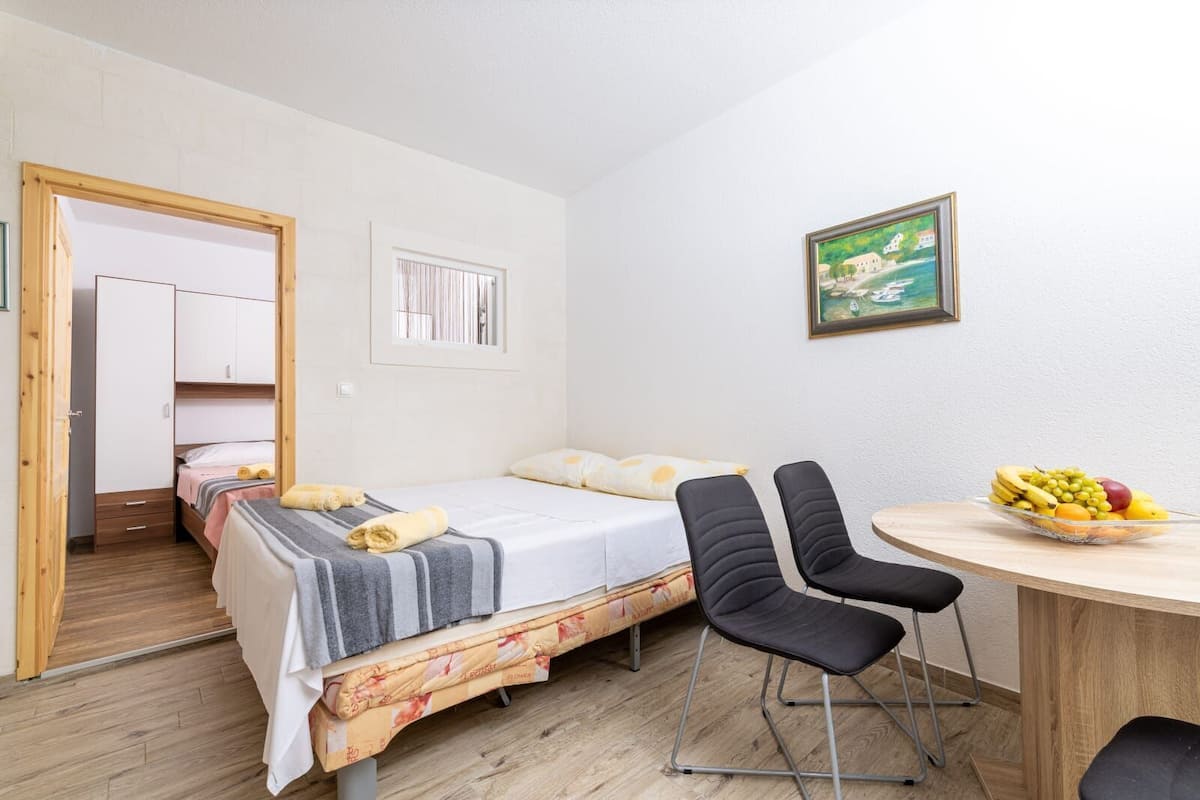 A small, well-lit room with two single beds, a wooden door, a small window, a framed painting, and a table with fruit.