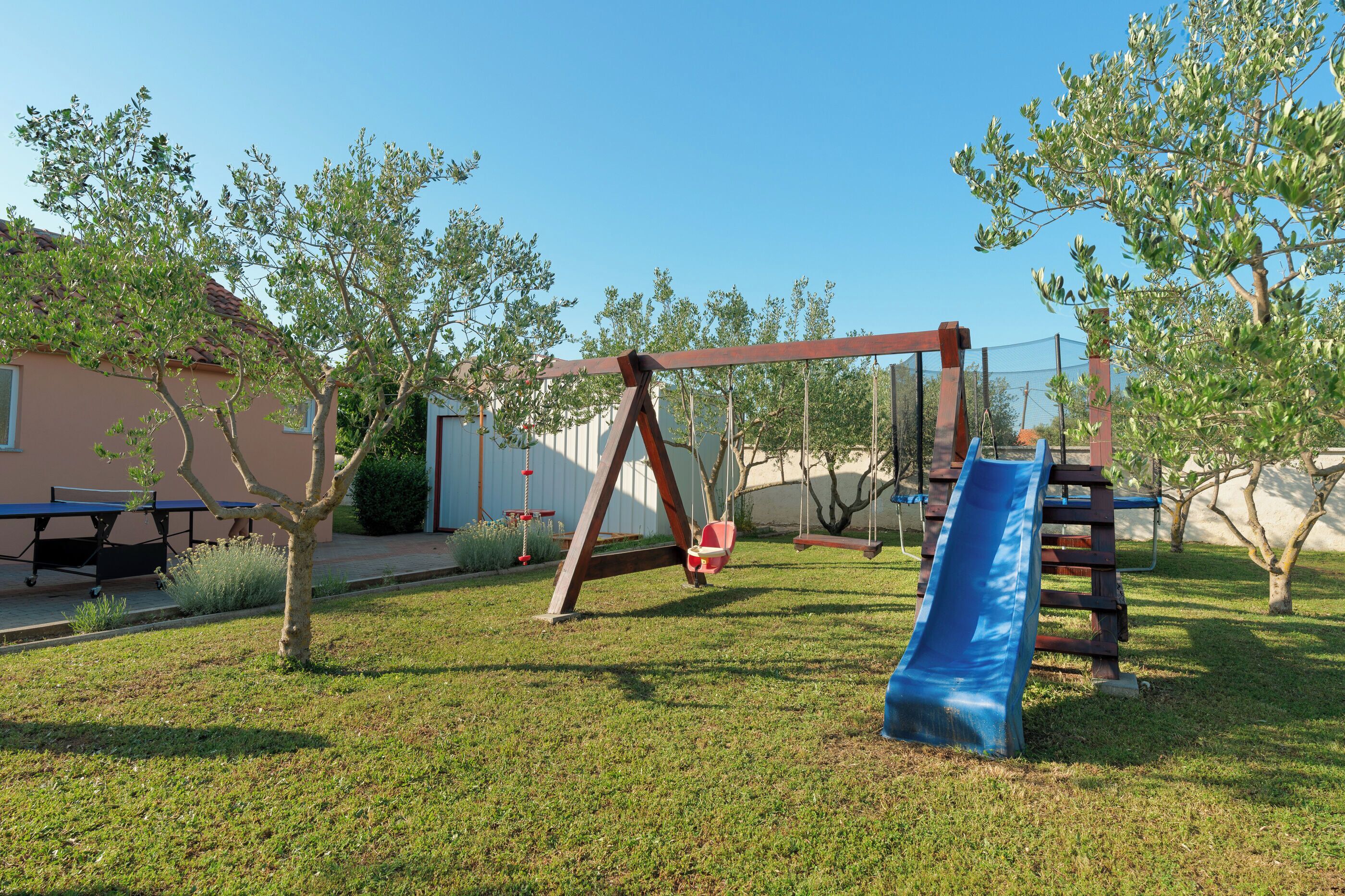 A playground with swings and a slide, surrounded by grass and trees.