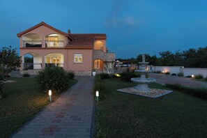 Una casa a due piani con facciata in pietra, un balcone e un giardino ben illuminato con una fontana.