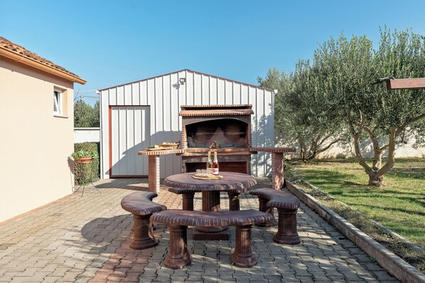Une terrasse extérieure avec une table et des bancs en briques, un barbecue et un bâtiment blanc en arrière-plan.