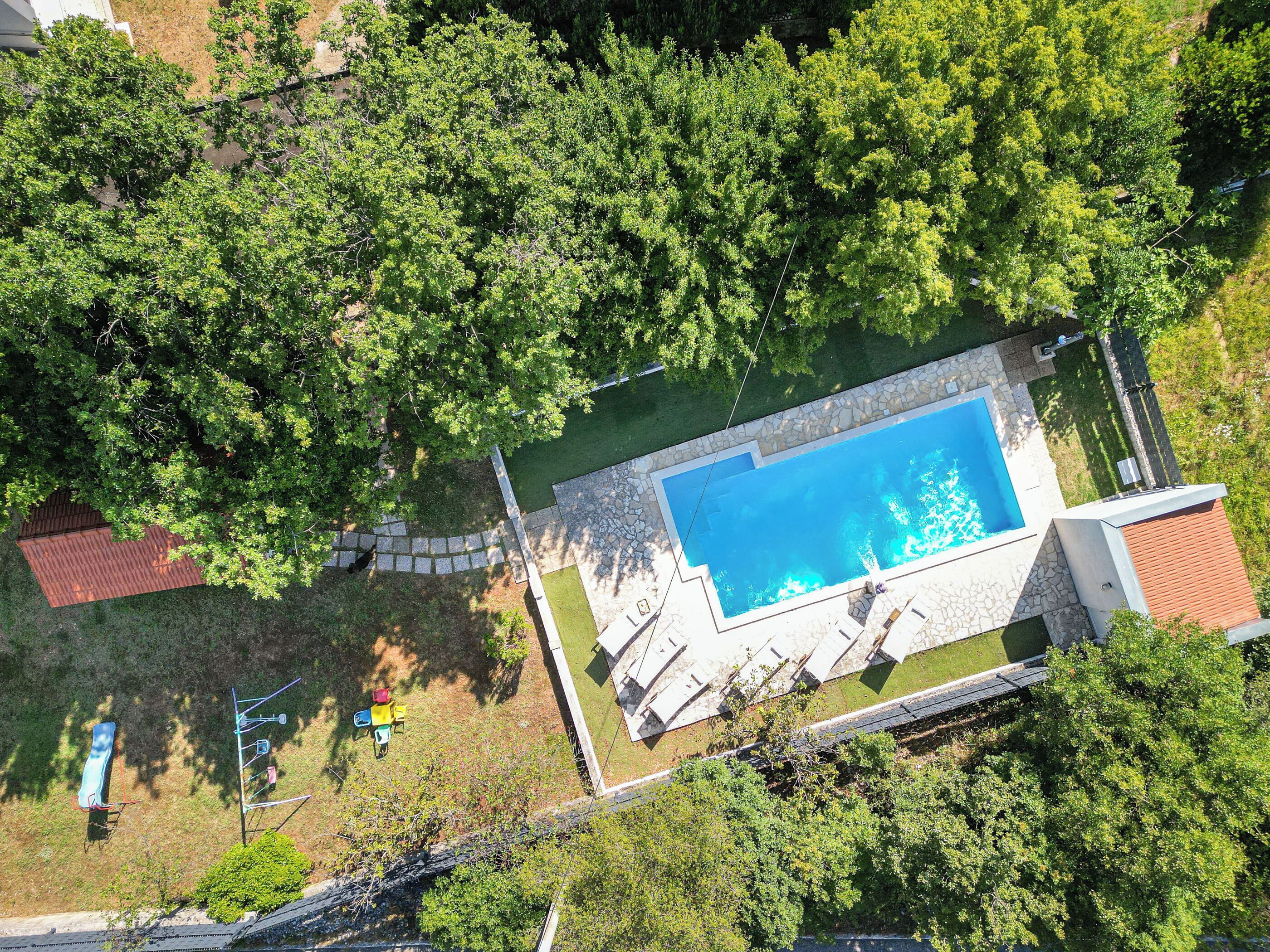 An aerial view of a residential property with a swimming pool surrounded by lush greenery, a stone pathway, and a small detached building.