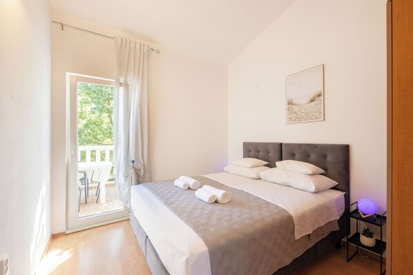 Ein Schlafzimmer mit einem Bett, einem Nachttisch mit Lampe, einem Fenster mit Vorhängen und Blick ins Freie.