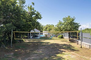 Ein umzäunter Garten mit Volleyballnetz, einer Terrassenfläche und einem Pool im Hintergrund.